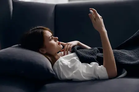Женщина лежит на диване с мобильным телефоном в руке, вид сбоку