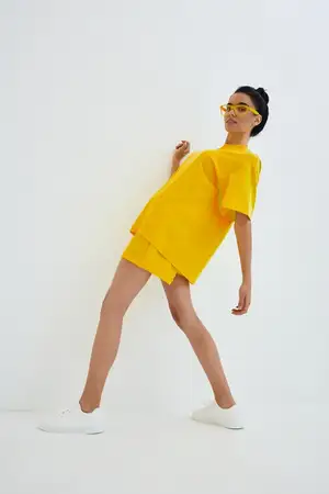 Стильная женщина, одетая в яркую желтую одежду oversized и солнцезащитные очки, динамично позирует с наклоном в минималистичном белом студийном пространстве