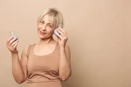 Счастливая женщина средних лет с блондинистыми волосами в бежевом топе держит две декоративные стеклянные вазы близко к лицу, мягко улыбаясь на однотонном бежевом фоне