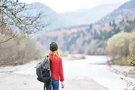 Счастливая женщина с рюкзаком отдыхает в горах у реки на фоне осеннего пейзажа, наслаждаясь свежим воздухом