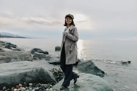 Улыбающаяся женщина в длинном пальто и берете на каменистом пляже у воды
