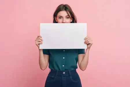 Молодая женщина с дружелюбной улыбкой стоит на розовом фоне, закрывая лицо пустой белой табличкой