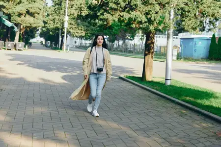 Повседневная уличная сцена: путешественница с фотоаппаратом идет по мощёной дорожке в парке среди деревьев и солнечного света