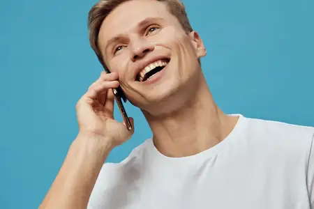 Счастливый улыбающийся мужчина пользуется телефоном, разговаривает по мобильному устройству, изолирован на голубом фоне, жизнерадостный молодой мужчина европеоидной расы