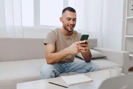 Улыбающийся мужчина взаимодействует со своим смартфоном, сидя на уютном диване в окружении светлого и просторного жилого пространства