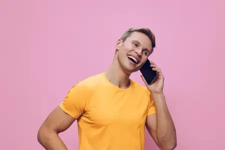 Счастливый молодой светлокожий мужчина улыбается, разговаривает по смартфону, одетый в повседневную оранжевую футболку, рука на бедре, стоит на розовом фоне
