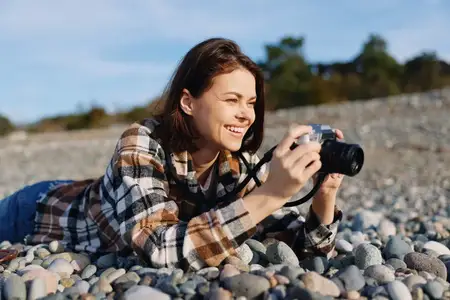 Повседневный фотограф на открытом воздухе отдыхает на каменистом берегу, с фотоаппаратом в руке и яркой улыбкой