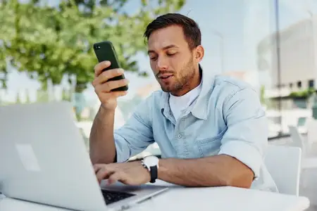 Молодой человек работает за ноутбуком, одновременно проверяя свой смартфон, сидя за столиком в уличном кафе