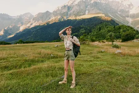 Женщина с рюкзаком на широком лугу на фоне возвышающихся вдалеке вершин, что наводит на мысли об исследованиях, свободе и связи с природой в условиях активного отдыха