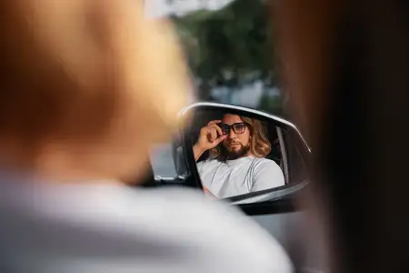 Задумчивый мужчина с длинными волосами и в очках всматривается в боковое зеркало, в котором отражается безмятежный уличный пейзаж