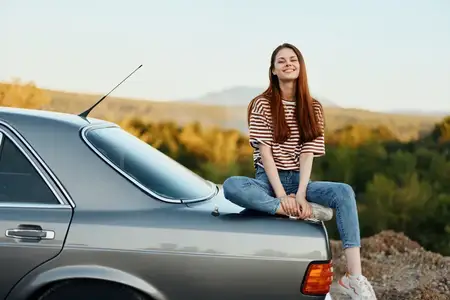 Женщина-водитель сидит на багажнике автомобиля и улыбается, любуясь красивым видом осенней природы и гор