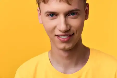 Улыбающийся молодой человек в желтой футболке в яркой студийной обстановке, теплые тона и расслабленная поза, повседневная мода, яркий фон, естественное освещение, минималистичная композиция