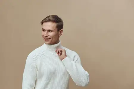 Весёлый молодой человек с короткими каштановыми волосами и сияющей улыбкой, одетый в уютный белый вязаный свитер, смотрит прямо в камеру, держа в руке мобильный телефон на фоне однотонного бежевого…