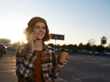 Городская женщина с чашкой кофе и телефоном, тёплая улыбка в шапке-бини и фланелевой рубашке, подсвеченная золотистым светом заката