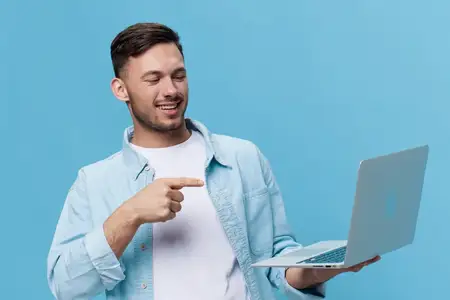 Улыбающийся счастливый загорелый красивый мужчина в повседневной базовой футболке указывает пальцем на новый купленный ноутбук и позирует изолированно на синем студийном фоне