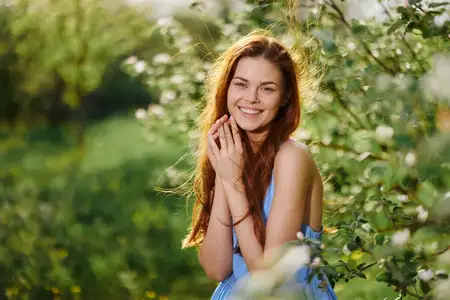 Весной женщина с развевающимися волосами стоит в саду цветущей яблони, улыбается, обнажая зубы, обнимает и гладит себя руками