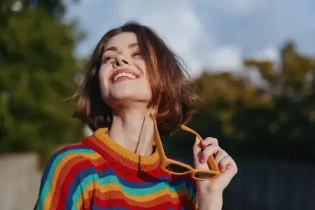Женщина улыбается в солнечных очках и радужном свитере, портрет на улице
