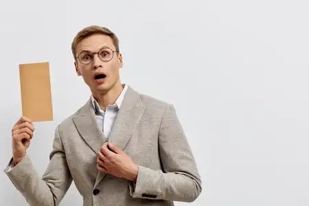 Молодой профессиональный мужчина, одетый в стиле smart casual, выражает удивление и любопытство, держа в руках пустую бежевую карточку на фоне однотонного серого студийного фона