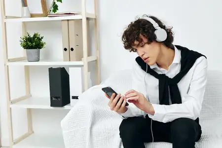 Кудрявый парень сидит на белом диване и смотрит в смартфон, листая социальные сети