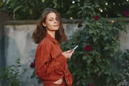 На ней ржавая рубашка и подходящие брюки у цветущей стены, залитой мягким дневным светом, что создаёт расслабленный жизненный момент с современным стилем и природным окружением