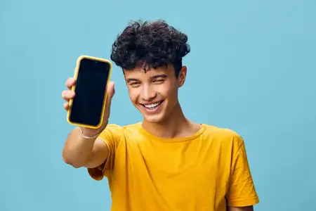 Веселый молодой человек в яркой желтой футболке держит смартфон с пустым экраном на фоне мягкого голубого фона