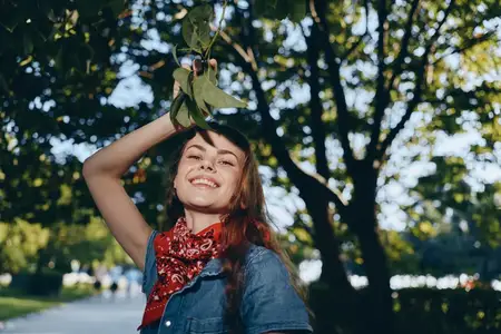 Улыбающаяся женщина, одетая в бохо-вестерн деним и красную бандану, стоит под густыми деревьями в теплом солнечном свете, запечатлевая беззаботный момент лайфстайла с эстетикой цветопередачи плёнки