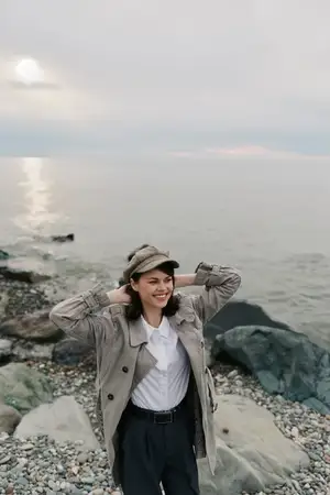 Счастливая молодая женщина в модном пальто и кепке стоит на галечном пляже с каменистым берегом и спокойным океаном на заднем плане