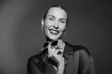 Портрет счастливой светлокожей женщины-европейки, позирующей в темной прозрачной блузе с солнцезащитными очками и серьгами-кольцами