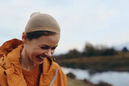 Женщина на открытом воздухе: смеется с опущенным взглядом в вязаной шапке и куртке у озера, зимний лайфстайл, яркий дневной свет, легкий ветерок, уютное настроение, живописный пейзаж в размытии