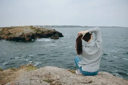 Женщина сидит у моря, держась за волосы, пейзаж, вид со спины