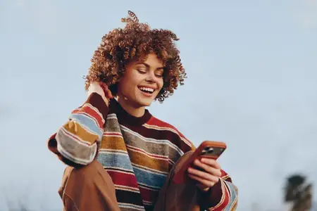 Женщина на улице, улыбается, пользуясь смартфоном, кудрявые волосы и повседневный полосатый свитер, общение в руке, солнечный свет, ощущение легкого ветерка, расслабленная поза, естественные цвета