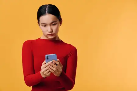 Женщина в красном топе держит смартфон, просматривая или печатая с серьезным лицом