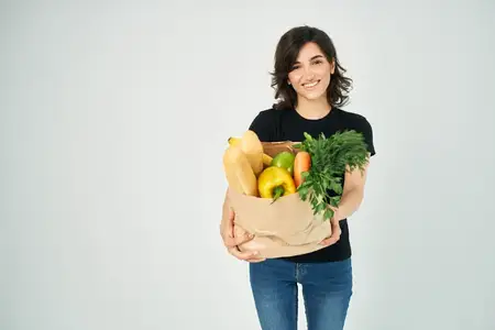 Весёлая женщина с пакетом продуктов, овощами, здоровой пищей на светлом фоне