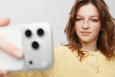 Улыбающаяся женщина с естественным макияжем и распущенными волосами держит телефон для селфи на простом белом фоне