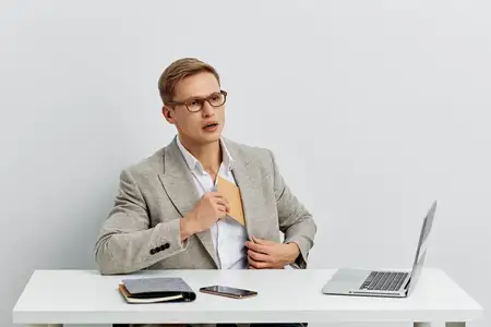 Молодой светлокожий мужчина работает как цифровой кочевник, сидя за белым столом с открытым ноутбуком и смартфоном, одетый в деловую повседневную одежду и очки, сосредоточен на удаленной работе и…