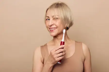 Счастливая женщина средних лет с короткими светлыми волосами улыбается, держа в руке электрическую зубную щетку на бежевом фоне