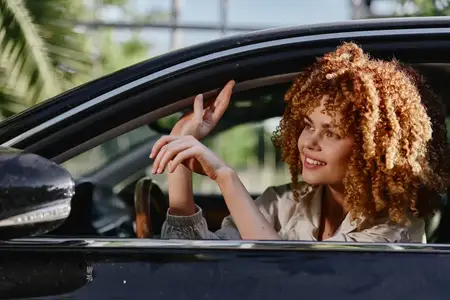 Улыбающаяся женщина с вьющимися волосами отдыхает у окна автомобиля, одетая в повседневную одежду, наслаждаясь ярким солнечным днём в окружении зелени, передавая беззаботное настроение