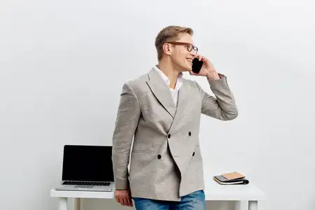 Улыбающийся мужчина разговаривает по смартфону, в современной серой куртке и повседневных джинсах, стоит рядом со столом с ноутбуком, цифровой кочевник, работающий удаленно с технологиями и связью…