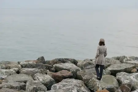Одинокая женщина в пальто идет по большим камням на морском побережье