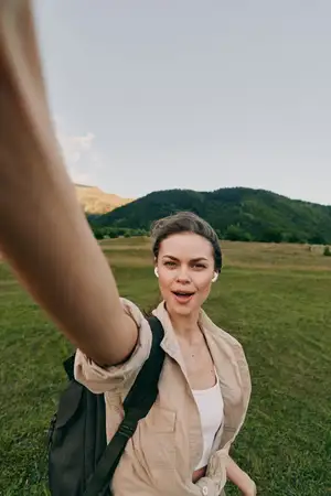 Счастливая женщина делает селфи на природе, на ней бежевая рубашка и белый топ, она стоит на травянистом поле с холмами позади