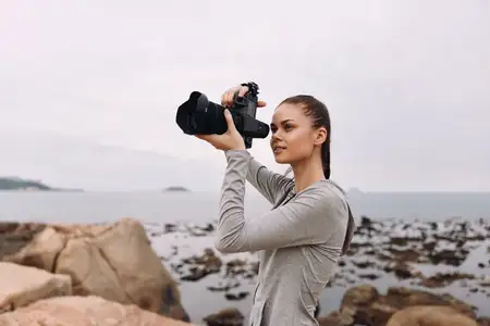 Целеустремленная молодая женщина в серой футболке с длинным рукавом фотографирует красивый прибрежный пейзаж, отражая свою любовь к фотографии в тихий день на пляже