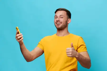 Привлекательный загорелый мужчина в жёлтой футболке с улыбкой показывает жест «палец вверх» и использует телефон, позируя изолированно на синем студийном фоне