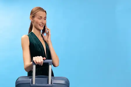 Улыбающаяся молодая женщина в зелёном платье с чемоданом разговаривает по телефону, изолирована на голубом фоне