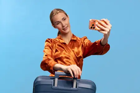 Улыбающаяся женщина с blond-волосами в оранжевой рубашке держит синий чемодан и делает селфи на смартфон на голубом фоне