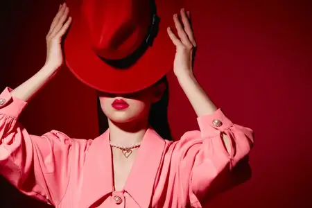 Молодая женщина в ярко-розовой блузке прячет лицо за огромной красной шляпой