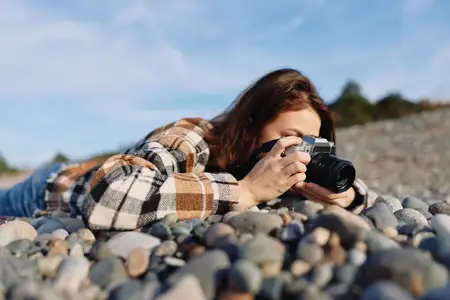 Сцена уличной фотосъёмки: сосредоточенная женщина на каменистом берегу, подняв камеру, в повседневной одежде