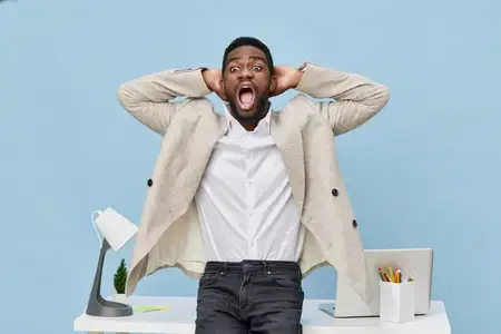 Взволнованный мужчина с юношеской внешностью, одетый в бежевый пиджак и белую рубашку, выражает шок или разочарование на рабочем месте, держа руки за головой, на фоне захламленного стола…