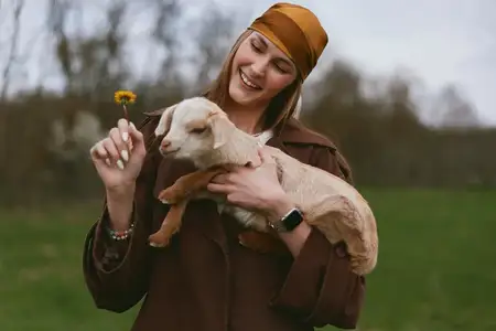 Молодая женщина в коричневом пальто и оранжевом платке нежно держит козленка и протягивает ему желтый цветок, стоя на травянистом поле с размытыми деревьями на заднем плане