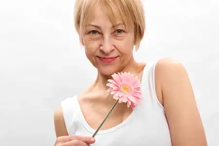 Пожилая женщина улыбается, цветок, розовый, весна, портрет, белый студийный фон, крупный план головы, когда она держит нежный цветок герберы, нежное выражение лица и мягкий свет создают спокойную…