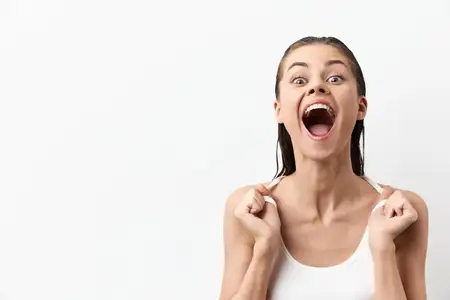Восторженная молодая женщина с мокрыми волосами в белой майке выражает счастье с открытым ртом и сияющими глазами на фоне чистого белого фона